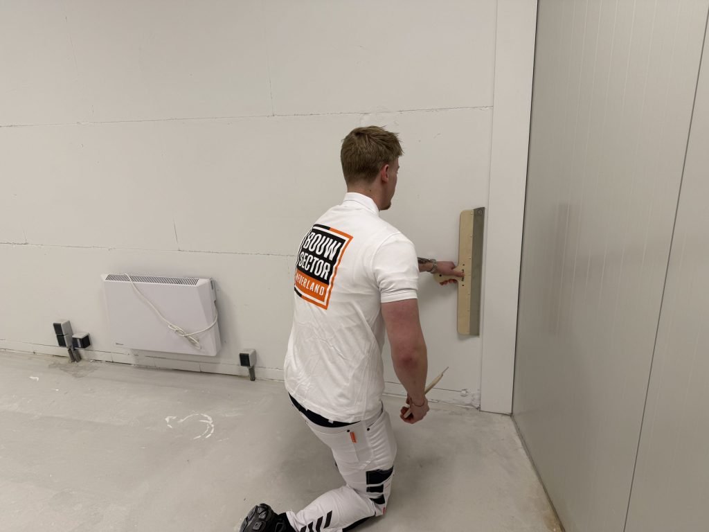 Stukadoor zit geknield en zet een lange spaan onderaan een wand bij de hoek strak